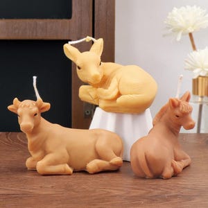 May include: Three animal-shaped wax candles in light brown and tan colors. The set includes a cow, a deer, and a horse, each in a resting position with a white wick protruding from the top. The figures feature detailed textures and facial features.