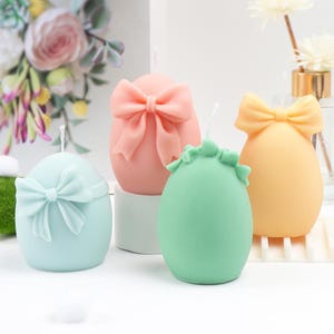 May include: Four pastel-colored egg-shaped candles with bow accents. The candles are in shades of light blue, pink, green, and yellow. Each candle has a wick at the top. Decorative candles for home decor.