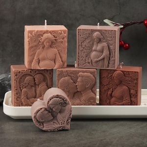 May include: A collection of six dusty rose-colored wax candles featuring intricate relief carvings of figures in various poses, including pregnant individuals and couples, with some square-shaped and one heart-shaped, all with a single wick.