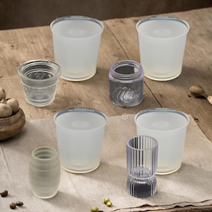 Op de afbeelding: Een set van zes heldere glazen bekers en potten in verschillende maten en vormen. De bekers zijn allemaal cilindrisch en hebben een gladde, matte afwerking. De potten hebben verschillende ontwerpen, waaronder een geribbeld patroon en een textuur. De bekers en potten zijn gerangschikt op een houten tafel met een lichtbruin tafelkleed.