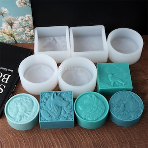 May include: A collection of soap molds and finished soaps. The molds are white and come in square, rectangular, and circular shapes. The finished soaps are teal and feature wolf designs, including a wolf howling at the moon. The soaps are arranged on a wooden surface.