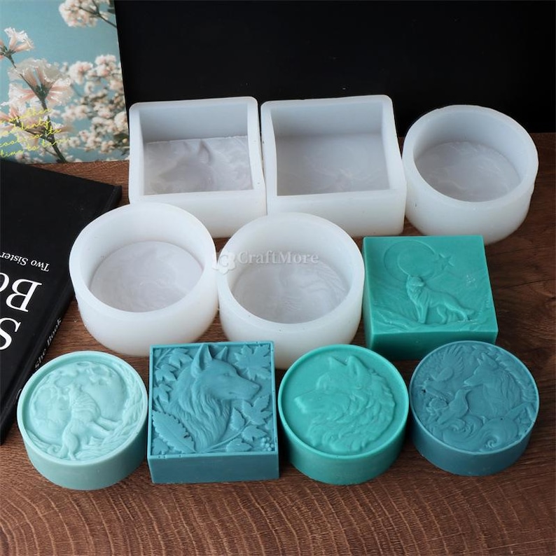 May include: A collection of soap molds and finished soaps. The molds are white and come in square, rectangular, and circular shapes. The finished soaps are teal and feature wolf designs, including a wolf howling at the moon. The soaps are arranged on a wooden surface.