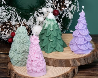 May include: Four decorative tree-shaped candles in shades of green, pink, and purple sit on wooden slices. The candles are intricately designed with floral and pine needle patterns. A festive wreath with pinecones and berries is in the background.