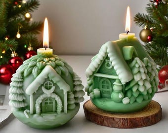 以下が含まれることがあります： 火の灯った芯のある、家を模した2つの緑色のクリスマスキャンドル。1つのキャンドルは木の台座に置かれています。キャンドルには、雪に覆われた屋根や装飾的な木など、複雑なディテールがあります。お祝いの休日の装飾。