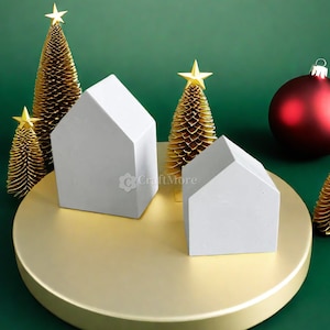 May include: Two white miniature houses with gold star-topped trees on a gold round platform. A red Christmas bauble is in the background.