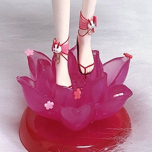 May include: A pink and white doll with white legs and feet wearing pink sandals with white bunny charms. The doll is standing on a pink lotus flower base.