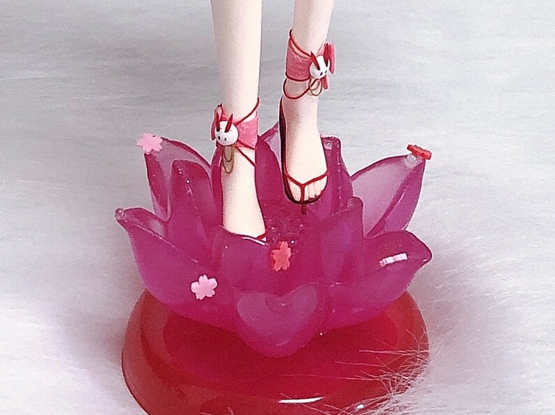 May include: A pink and white doll with white legs and feet wearing pink sandals with white bunny charms. The doll is standing on a pink lotus flower base.