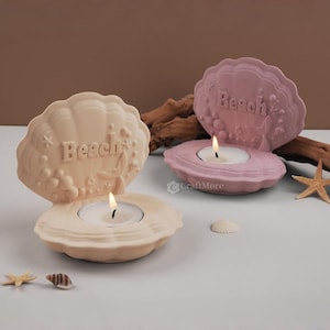Puede incluir: Dos portavelas de cerámica con forma de concha marina con la palabra "Beach" escrita en ellos. Los portavelas son de color rosa y beige y tienen una pequeña vela de té en su interior.