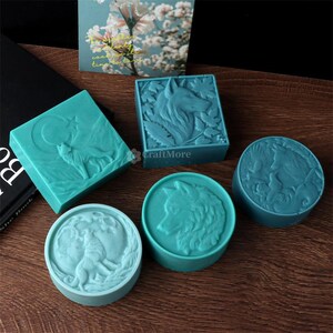 May include: A collection of teal-colored soaps with wolf designs. The soaps are square and round, with detailed relief carvings of wolves, moons, and foliage. The soaps are arranged on a wooden surface, with a floral card in the background.