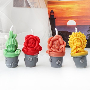 May include: Four small, hand-crafted, scented candles shaped like potted plants. Each candle features a baby face with a different flower design: a cactus, a red rose, a pink rose, and a sunflower.
