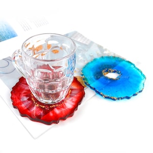 May include: Two resin coasters with a geode design. The coaster on the left is red with gold flecks and the coaster on the right is blue with gold flecks. A clear glass mug is sitting on the red coaster.