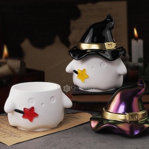 Könnte beinhalten: Zwei weiße Keramik-Geisterfiguren mit schwarzen und goldenen Hexenhüten. Ein Geist trägt einen lila und goldenen Hexenhut. Die Geister halten einen gelben Stern und einen roten Stern.