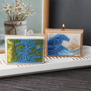May include: Two rectangular candles with a light brown base and a colorful image on the front. The candle on the left has a blue and yellow image of a starry night scene. The candle on the right has a blue and white image of a large wave. The candle on the right has a lit wick.