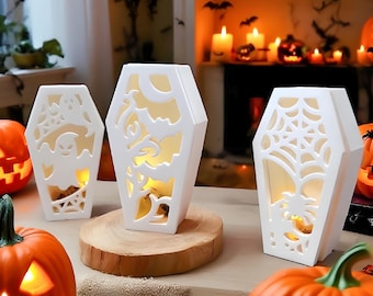Puede incluir: Tres faroles blancos con forma de ataúd para Halloween con diseños recortados de un fantasma, murciélagos y una telaraña. Los faroles están iluminados desde dentro, colocados sobre una superficie de madera con calabazas talladas cerca.