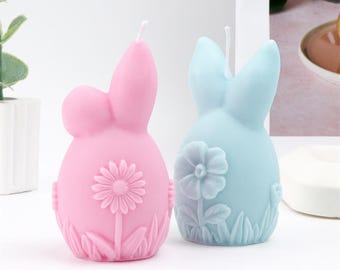 May include: Two bunny-shaped candles. One is pink, the other is light blue. Each candle has a flower design on the front and a wick at the top. The candles are decorative and suitable for Easter.