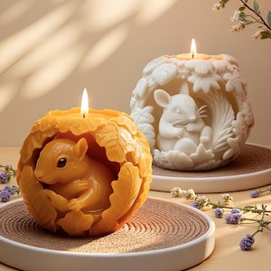 May include: Two decorative candles, one orange and one white, each featuring a woodland animal design. The orange candle has a squirrel inside a leaf-shaped exterior. The white candle features a mouse nestled in a leafy design. Both candles are lit.