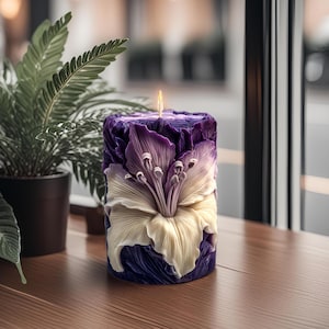 May include: A cylindrical candle with a floral design. The candle is primarily purple with a cream-colored flower in the center. The candle is lit, with a small flame at the top. The candle is on a wooden surface.