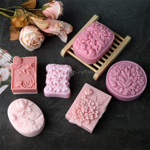 Peut inclure: Divers savons roses avec des motifs floraux et botaniques. Les savons présentent des formes variées, rectangulaires, ovales et rondes, avec des sculptures détaillées de roses, de fleurs et de feuilles. Un savon est posé sur un petit porte-savon en bois.