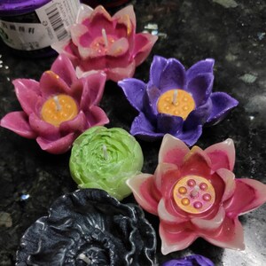 May include: Assortment of colorful lotus flower shaped candles. The candles are in shades of pink, purple, green, and black. Each candle has a small wick in the center and a decorative design.