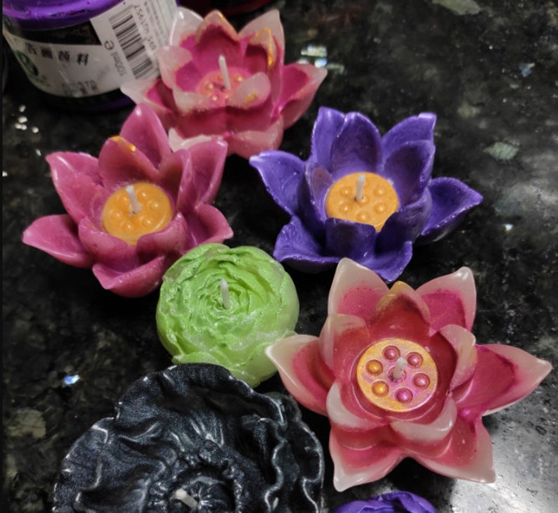May include: Assortment of colorful lotus flower shaped candles. The candles are in shades of pink, purple, green, and black. Each candle has a small wick in the center and a decorative design.