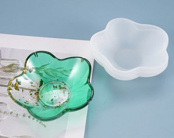 May include: A green glass bowl with gold flecks and a flower-shaped rim. The bowl is sitting on a white surface next to a clear silicone mold with a similar flower-shaped rim.
