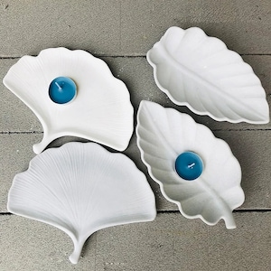 May include: Four white ceramic leaf-shaped dishes with raised veins. Each dish has a small blue tealight candle in the center.
