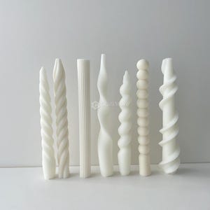 May include: A selection of white decorative candles in diverse forms. The candles feature twisted, spiral, and column designs. The candles are arranged on a white surface against a white backdrop. The candles are unscented and are for decorative use.