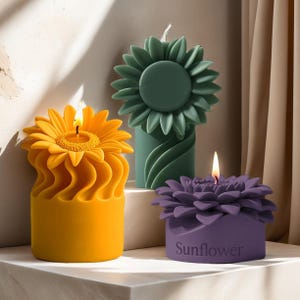 Op de afbeelding: Drie zonnebloemvormige kaarsen in geel, groen en paars. De gele kaars is aangestoken en toont een heldere vlam. De paarse kaars heeft het woord "Sunflower" op de basis. De kaarsen staan op een wit oppervlak.