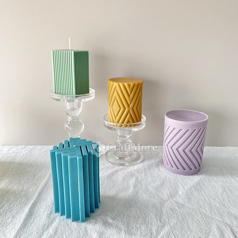 May include: Three decorative pillar candles with geometric patterns. The candles are green, yellow, and purple. The candles are on a white surface.