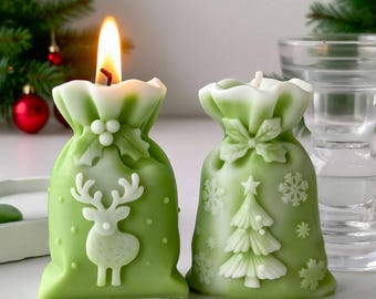 May include: Two green Christmas-themed candles shaped like gift bags. One candle has a deer design, the other a Christmas tree and snowflakes. The candles are lit, with a white and green color scheme.