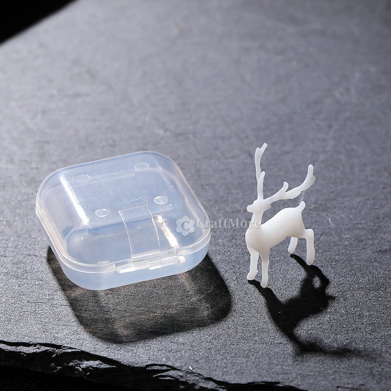 Pu&ograve; includere: Una piccola statuina di cervo bianca con corna, in piedi su una superficie grigia. Nell'immagine &egrave; presente anche un piccolo contenitore quadrato di plastica trasparente.