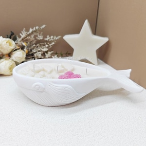 May include: A white whale-shaped candle holder filled with white wax, decorative stones, and a pink flower. A white star-shaped candle is in the background. The candle holder has a textured surface and is on a white surface.