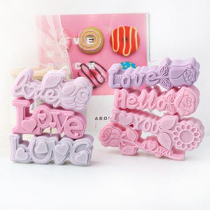 Peut inclure: Deux ensembles de bougies empilées avec des messages différents. Le premier ensemble épelle "LOVE" trois fois en violet et rose. Le deuxième ensemble épelle "Love", "Hello", "I Love You" et "I Love You" en rose et violet.