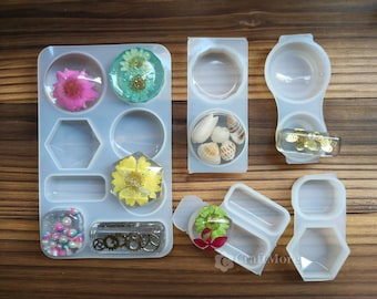 May include: A collection of clear silicone molds in various shapes, including circles, hexagons, and rectangles. Some molds contain dried flowers, seashells, and small gears. The molds are arranged on a wooden surface.