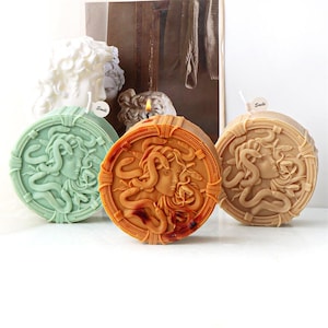 May include: Three round candles with a Medusa design in shades of green, orange, and beige. The candles have a detailed relief of Medusa's face and snakes. A white sculpture of a bust is in the background.