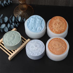 May include: A collection of round, cat-themed soap molds in various colors. The molds feature detailed cat faces and cat pairs, with heart and floral accents. The soaps are displayed on a wooden rack and a dark surface.