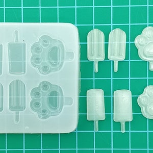 May include: A clear silicone mold with 6 cavities for making ice pops and paw print shapes. The mold is on a green grid background.