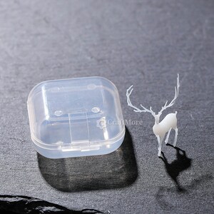 Pu&ograve; includere: Una scatola di contenimento in plastica trasparente con un coperchio incernierato e una statuetta di cervo bianca con corna.