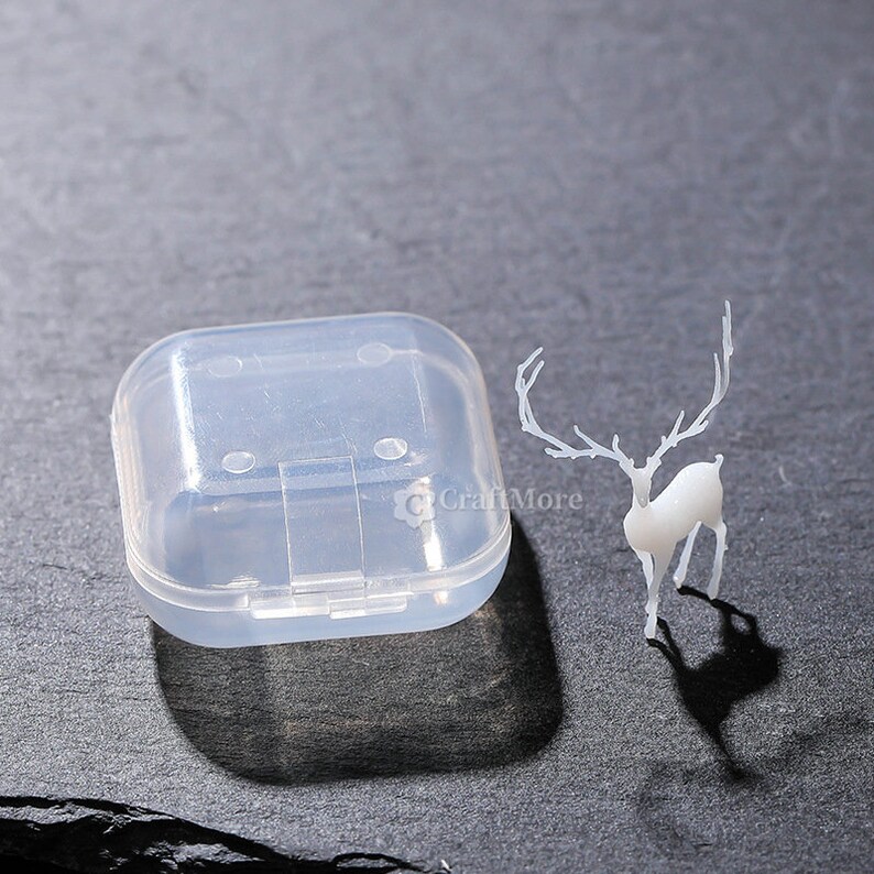 Pu&ograve; includere: Una scatola di contenimento in plastica trasparente con un coperchio incernierato e una statuetta di cervo bianca con corna.
