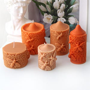 May include: Four orange and brown pillar candles with a honeycomb and bee design. The candles are arranged in a row on a white surface.