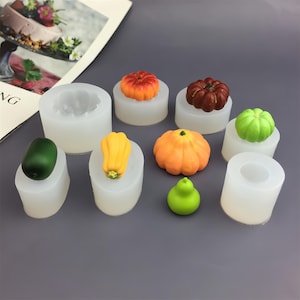 May include: A selection of miniature pumpkins and gourds in various colours, including orange, red, yellow, and green. They are displayed on small, white, cylindrical moulds. The image evokes a harvest or autumn theme.