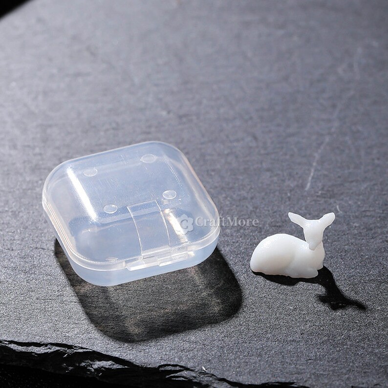 Pu&ograve; includere: Una piccola statuina di cervo bianca, seduta su una superficie grigia, accanto a un contenitore di stoccaggio in plastica trasparente con un coperchio incernierato.