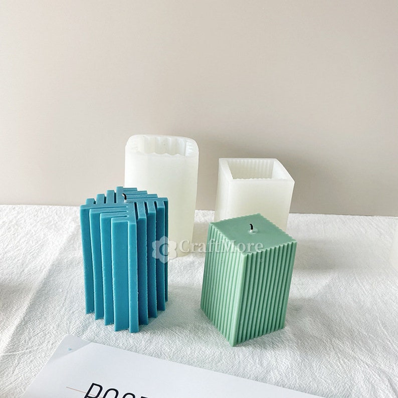 May include: Three candle moulds, two are white silicone and one is a teal blue geometric candle. The teal blue candle is in a geometric shape with a square base and a rectangular top. The white silicone moulds are square and rectangular.