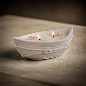 Könnte beinhalten: Ein weißer Kerzenhalter aus Keramik in Form eines Bootes mit zwei brennenden Kerzen und Muscheln im Inneren. Das Boot hat ein Seil-Design am Rand.