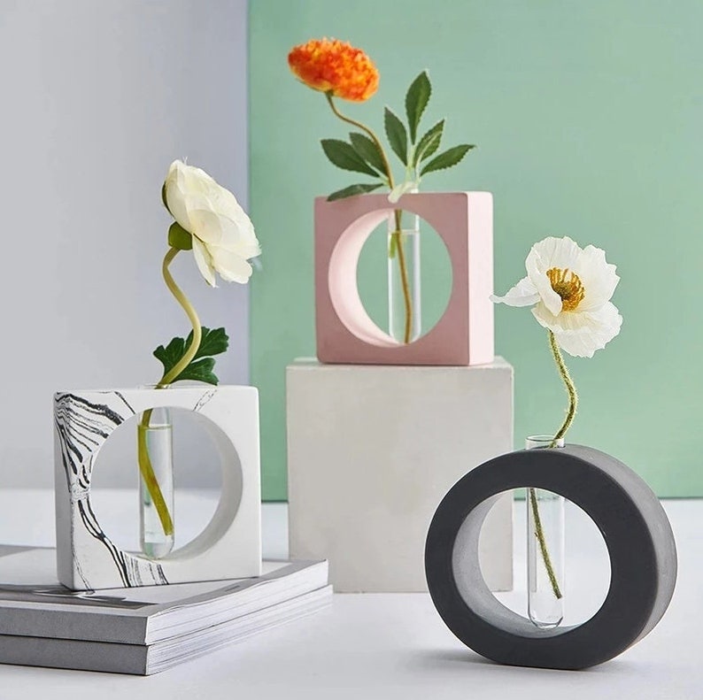 May include: Three modern concrete vases with glass tubes for holding flowers. The vases are in a square, round, and marble pattern. The flowers are white and orange.