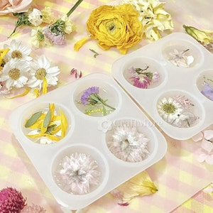 May include: A white silicone mold with four round cavities, each filled with pressed flowers and leaves. The mold is on a pink and white patterned surface.