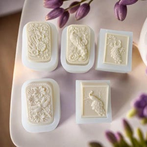 Può includere: Cinque stampi per sapone bianco sporco con disegni a rilievo dettagliati. Gli stampi presentano motivi floreali, un pavone e una libellula. I saponi sono inseriti in stampi rettangolari e quadrati bianchi, di colore morbido e cremoso. Vista dall'alto.