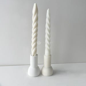 May include: Two white twisted taper candles in matching white holders. The candles have a spiral design and are of different heights. The holders have a rounded base and a cylindrical top. The background is a solid white.