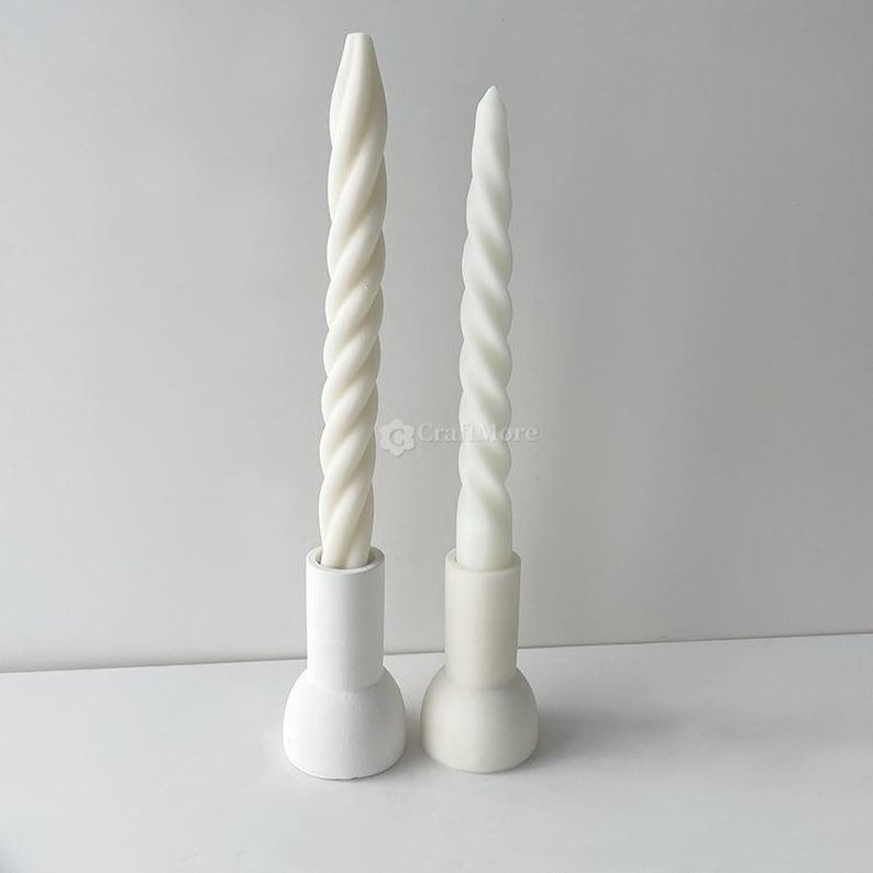 May include: Two white twisted taper candles in matching white holders. The candles have a spiral design and are of different heights. The holders have a rounded base and a cylindrical top. The background is a solid white.