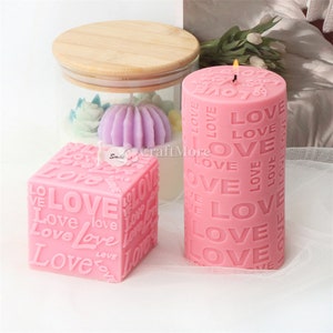 May include: Two pink candles with the word "LOVE" embossed on them. The candle on the left is a square shape and the candle on the right is a cylinder shape.